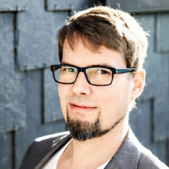 Christian, lächelnd, mit einer Brille mit dunklem Rahmen, braunen Augen und einem kurzen Bart.