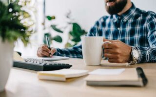 Nahaufnahme eines Mannes in einem karierten Hemd, der an einem Schreibtisch sitzt und in der einen Hand eine große weiße Tasse hält, während er mit der anderen Hand in ein Notizbuch schreibt. Auf dem Schreibtisch befinden sich eine Tastatur und ein Notizblock, im Hintergrund eine Topfpflanze und andere Büromaterialien.
