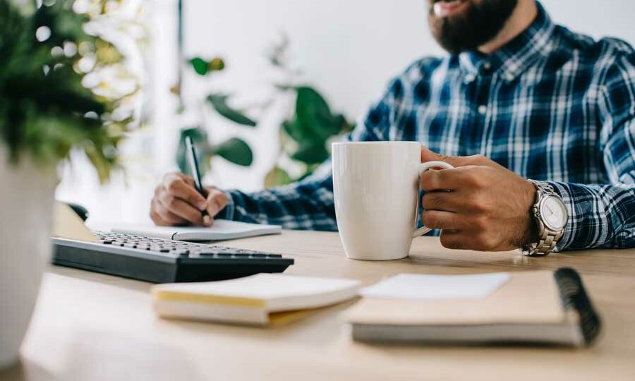 Nahaufnahme eines Mannes in einem karierten Hemd, der an einem Schreibtisch sitzt und in der einen Hand eine große weiße Tasse hält, während er mit der anderen Hand in ein Notizbuch schreibt. Auf dem Schreibtisch befinden sich eine Tastatur und ein Notizblock, im Hintergrund eine Topfpflanze und andere Büromaterialien.